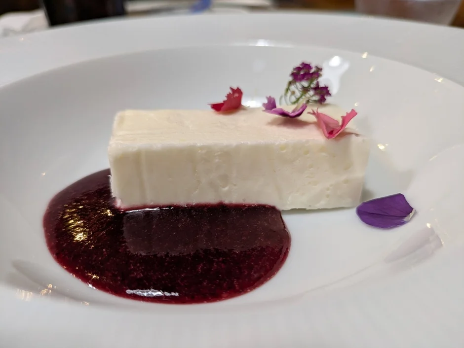 白い四角いデザートが白い皿に盛られ、鮮やかなベリーソースが下に敷かれています。ピンクや紫の花びらで上品に飾られた、美しい一皿のスイーツです。