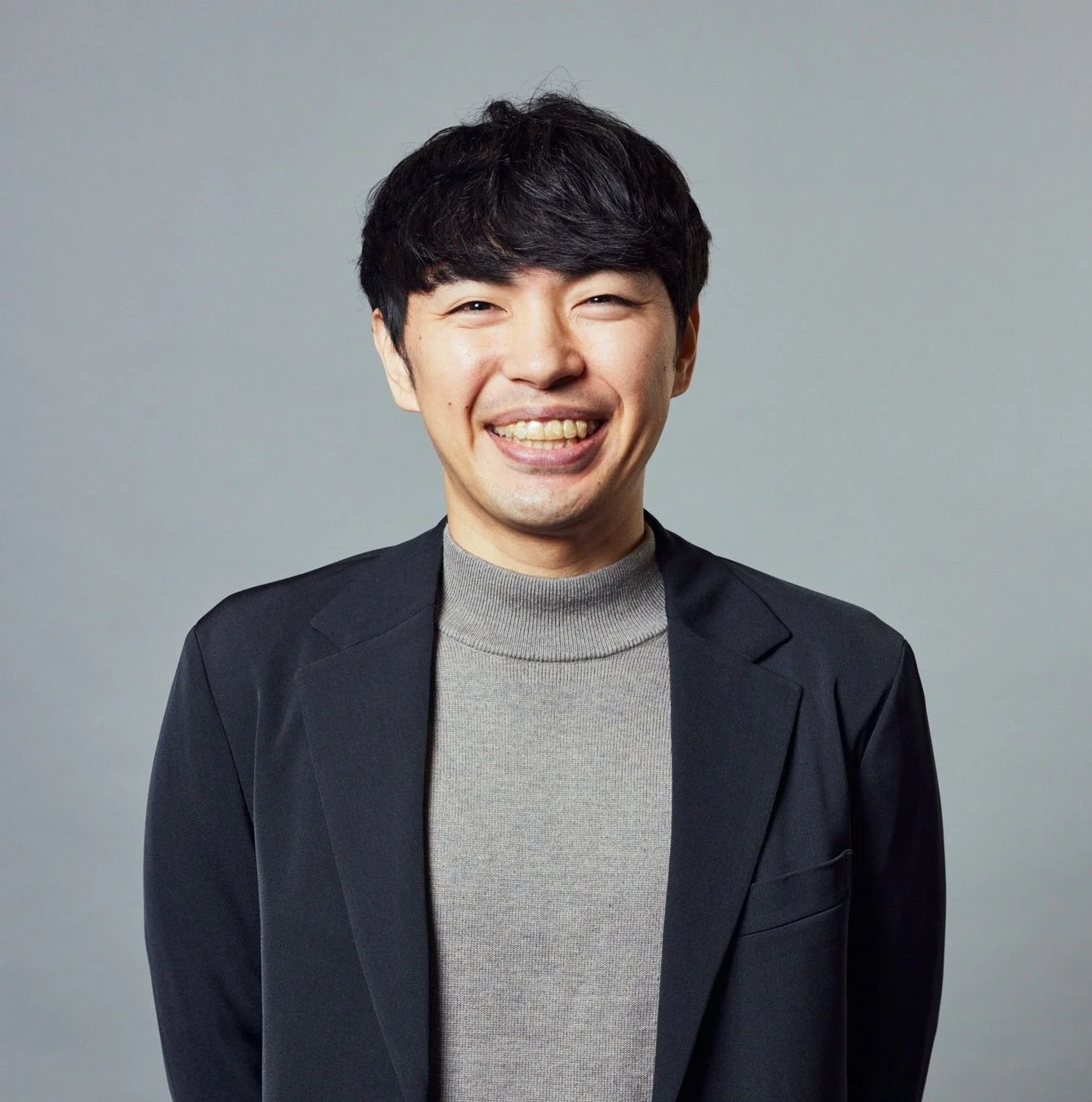 グレーのタートルネックと黒いジャケットを着用し、明るい笑顔でカメラを見つめるアジア人男性のポートレート。親しみやすくプロフェッショナルな印象です。