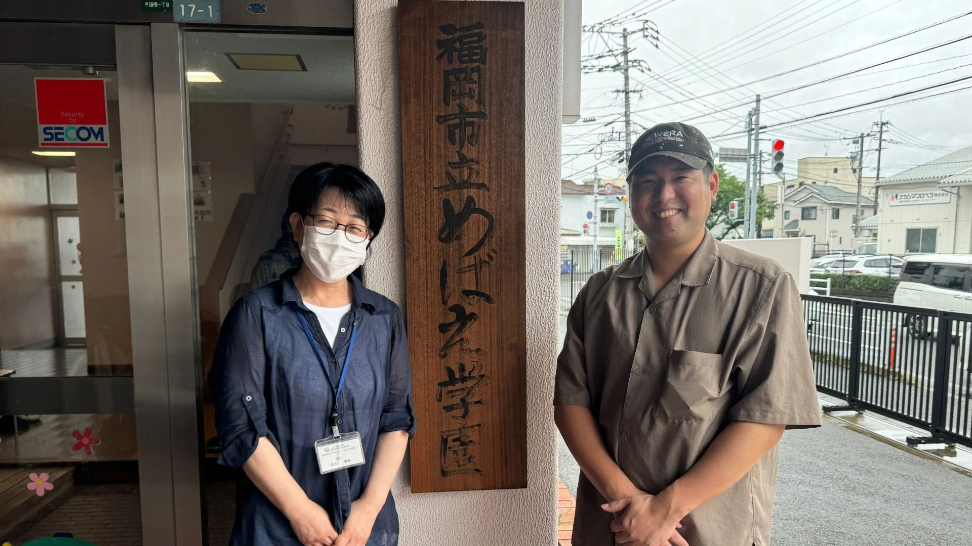 福岡市立めばえ学園の看板の前で、笑顔の男女2人が立っている写真です。