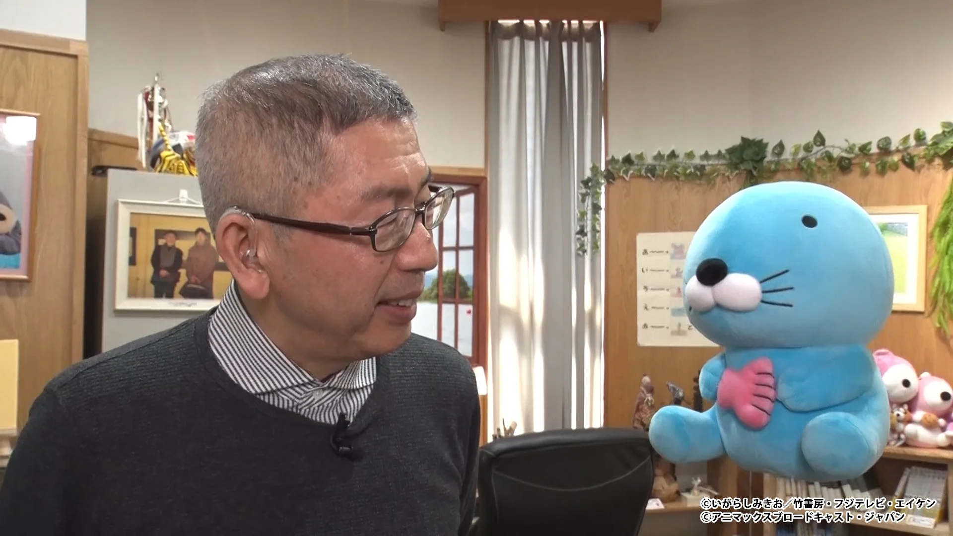 眼鏡をかけた男性が青いカワウソのようなキャラクターのぬいぐるみと並んで座っている