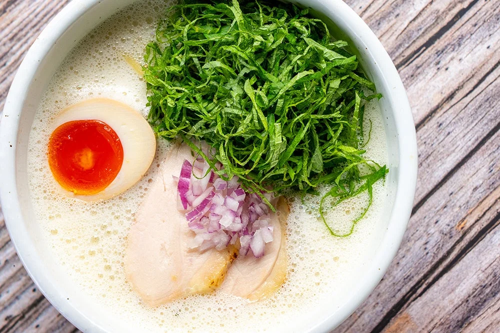 白い器に入ったクリーミーなスープのラーメン、半熟卵、薄切り鶏肉、刻み紫玉ねぎ、青ネギ