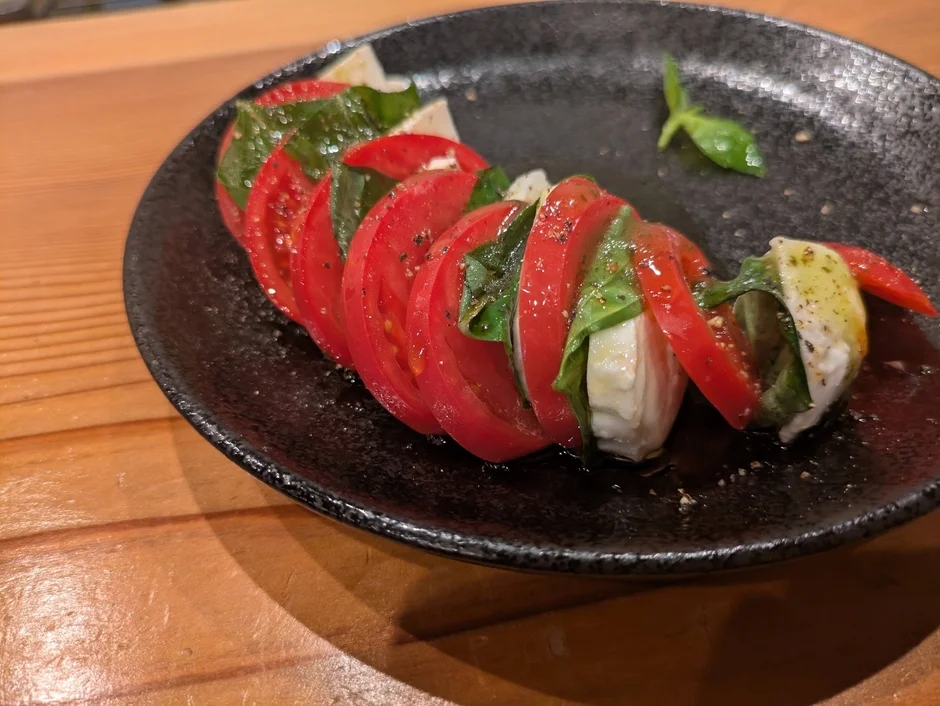 黒い皿に盛り付けられたカプレーゼサラダです。赤くスライスされたトマトと白いモッツァレラチーズ、緑のバジルが交互に並び、オリーブオイルと黒胡椒がかかっています。新鮮で彩り豊かな一品です。