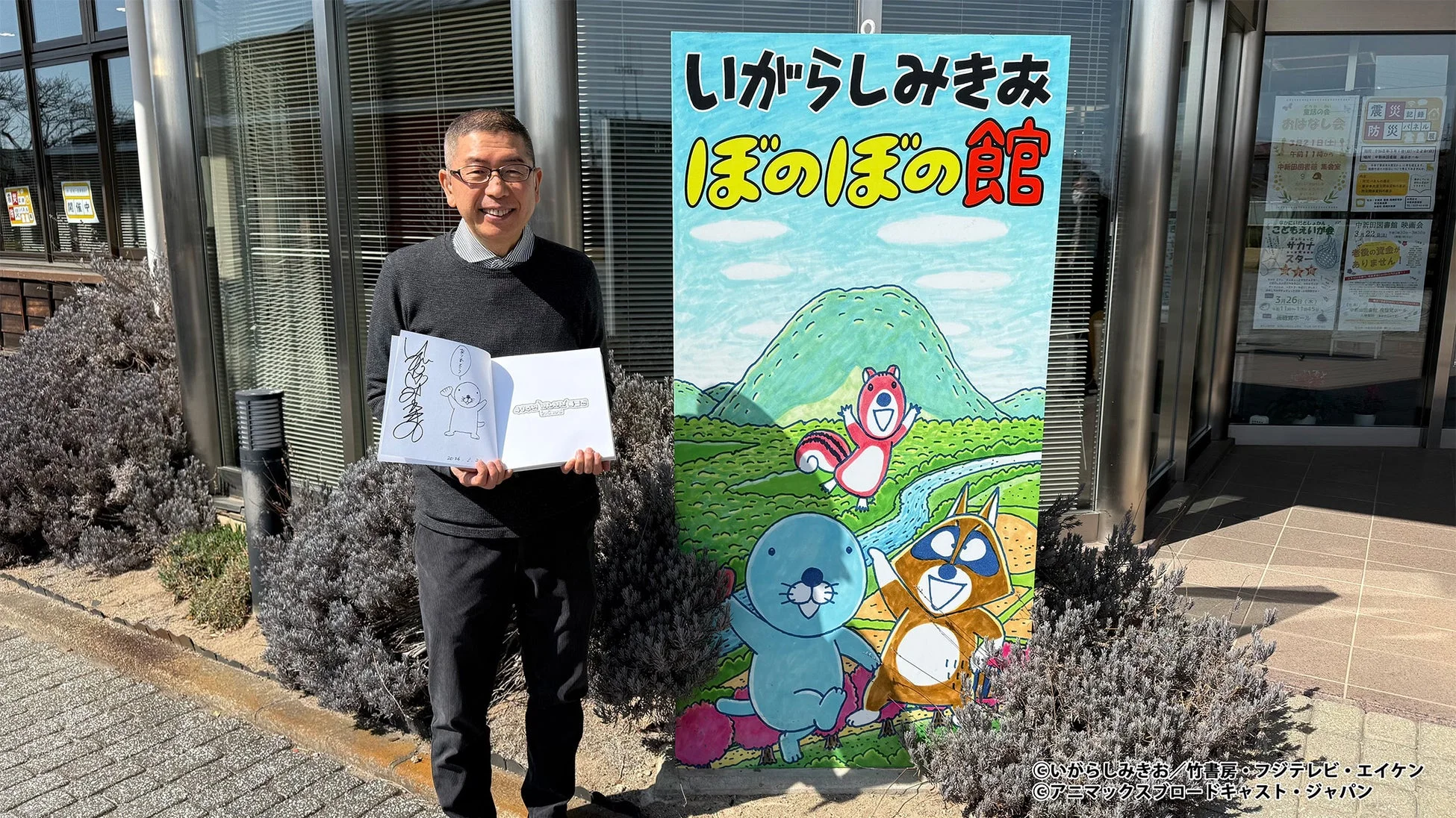 漫画家のいがらしみきお氏らしき男性が、自身の作品「ぼのぼの」のキャラクターが描かれた看板の前で、サイン入りの本を手に笑顔で立っている