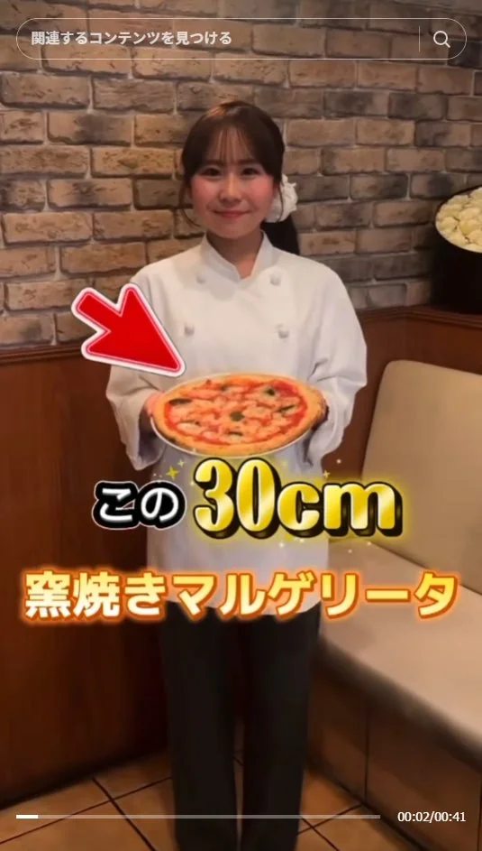 シェフの制服を着た女性が30cmの窯焼きマルゲリータピザを掲げている画像