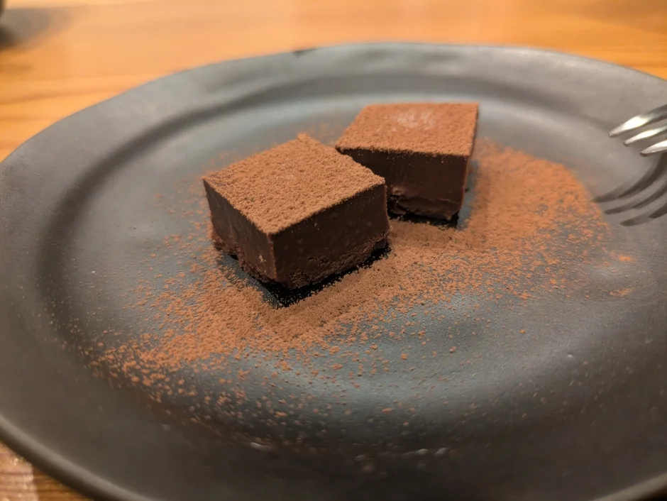 黒い皿に盛り付けられた2切れの生チョコレート。表面にはココアパウダーがまぶされ、皿の周りにも散らばっています。デザートとして提供されている様子で、フォークの一部も写っています。