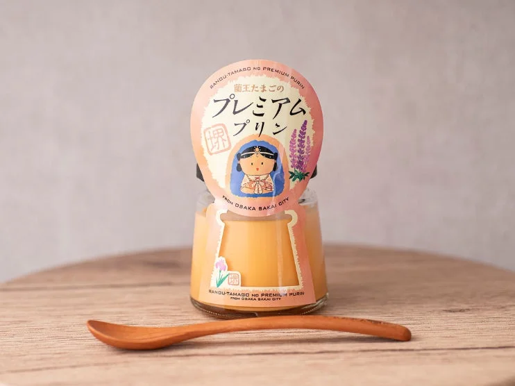 蘭王たまごを使用したプレミアムプリンが、木製スプーンと共に木目のテーブルに置かれている画像です。