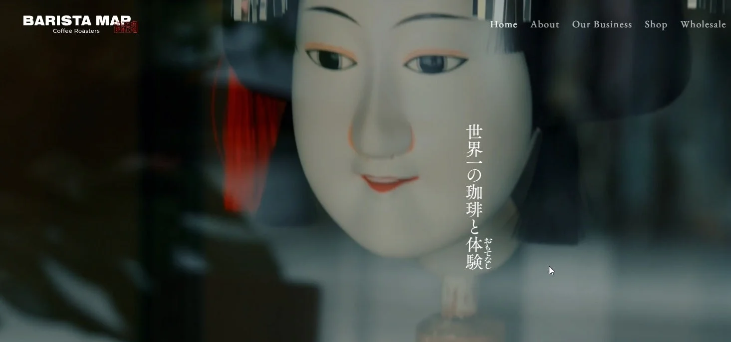 ウェブサイトのトップページと思われる画像で、中央に日本の伝統的な人形の顔が大きく写っています。右側には「世界一の珈琲と体験」「おもてなし」という日本語のテキストがあり、左上には「BARISTA MAP Coffee Roasters」というロゴとナビゲーションメニューが見えます。
