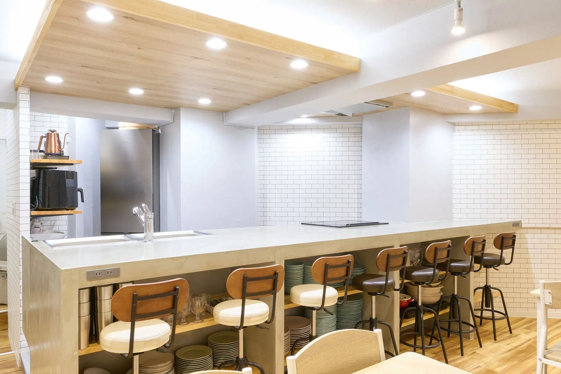 モダンで清潔感のあるキッチン兼ダイニングスペースです。長いコンクリート調のカウンターに複数のバースツールが並び、奥には冷蔵庫や調理器具が見えます。
