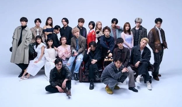 明るいスタジオで、多様なファッションに身を包んだ20人ほどの若い男女が集合写真を撮影している様子。様々なポーズで個性を表現している。