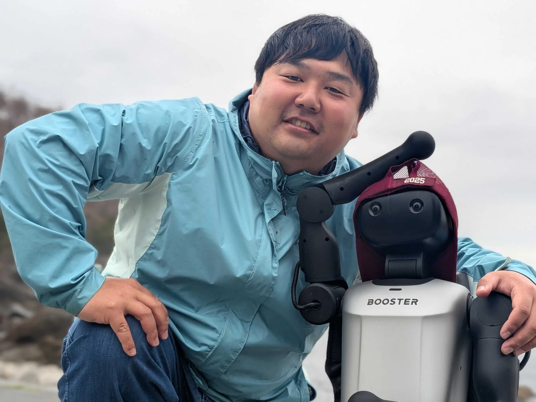 水色のジャケットを着た男性が、BOOSTERという名前のロボットと一緒に屋外で笑顔を見せています。ロボットは「2025」と書かれたエンジ色の帽子をかぶっています。