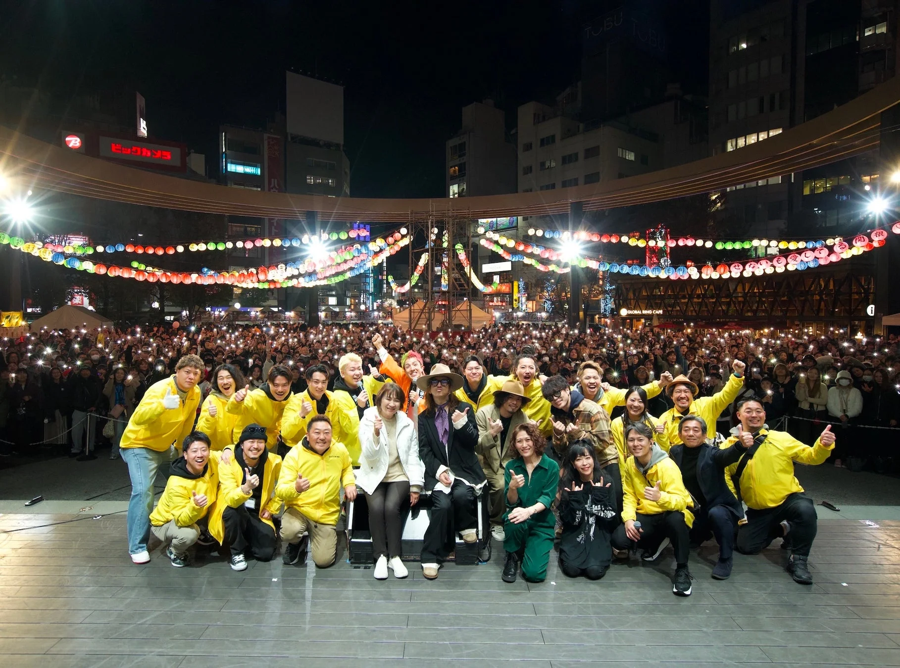 夜の広場で開催されたイベントで、ステージに立つ多くの出演者たちが観客をバックに集合写真を撮っている様子