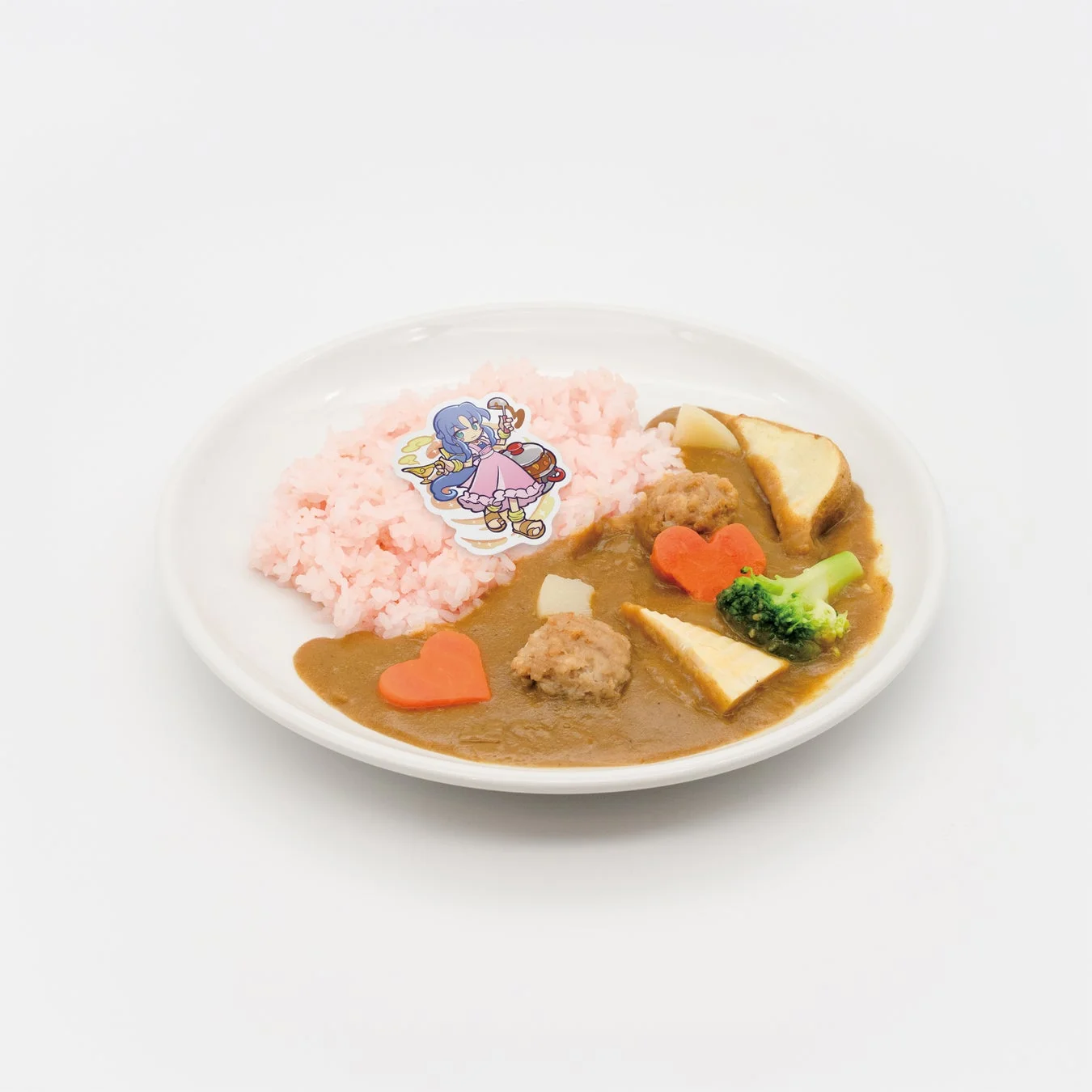 白い皿に盛られたピンク色のライスとカレー。カレーにはミートボール、ハート型の人参、ブロッコリー、ジャガイモ、パンが添えられ、ピンクのライスの上には可愛いアニメキャラクターのピックが飾られています。