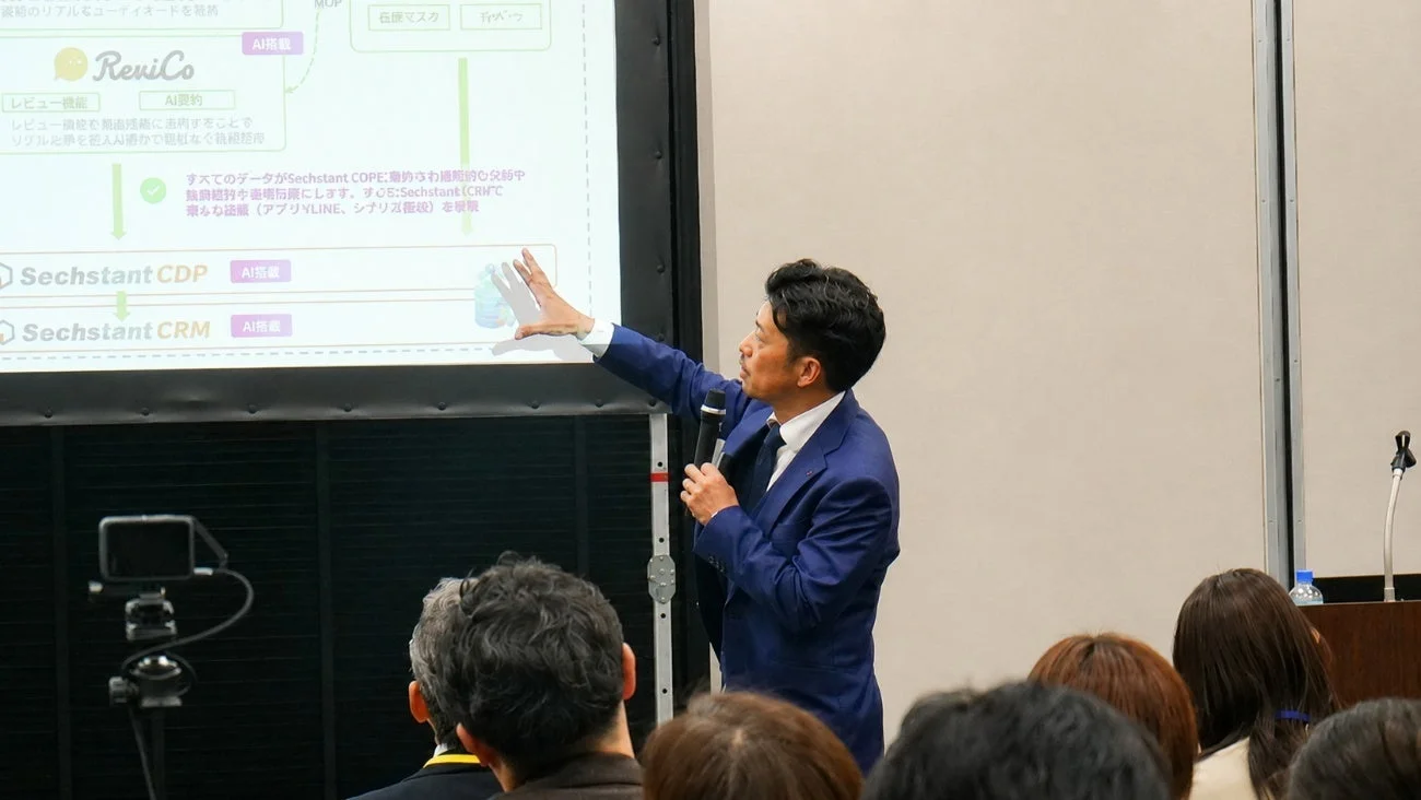 プレゼンテーションの様子。ビジネスシステムに関する日本語の図とテキストがスクリーンに表示されている