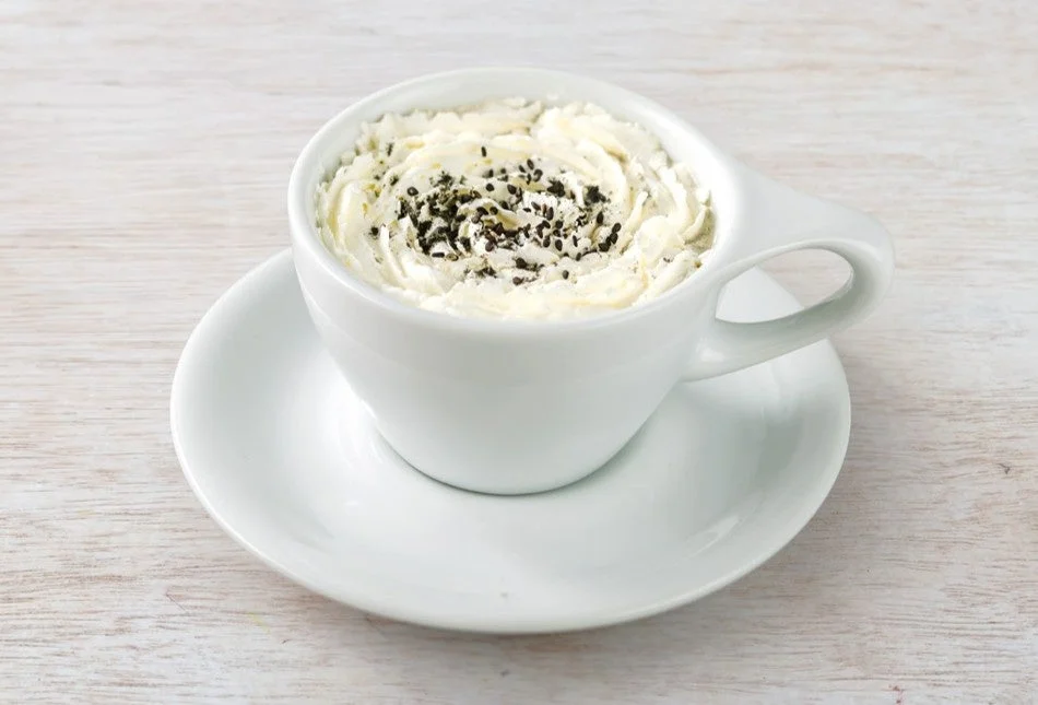 白い木目のテーブルに置かれた白いカップに入った飲み物。たっぷりのホイップクリームが渦巻き状に盛られ、黒ごまがトッピングされている。温かいカフェドリンクのようで、リラックスした雰囲気を演出しています。