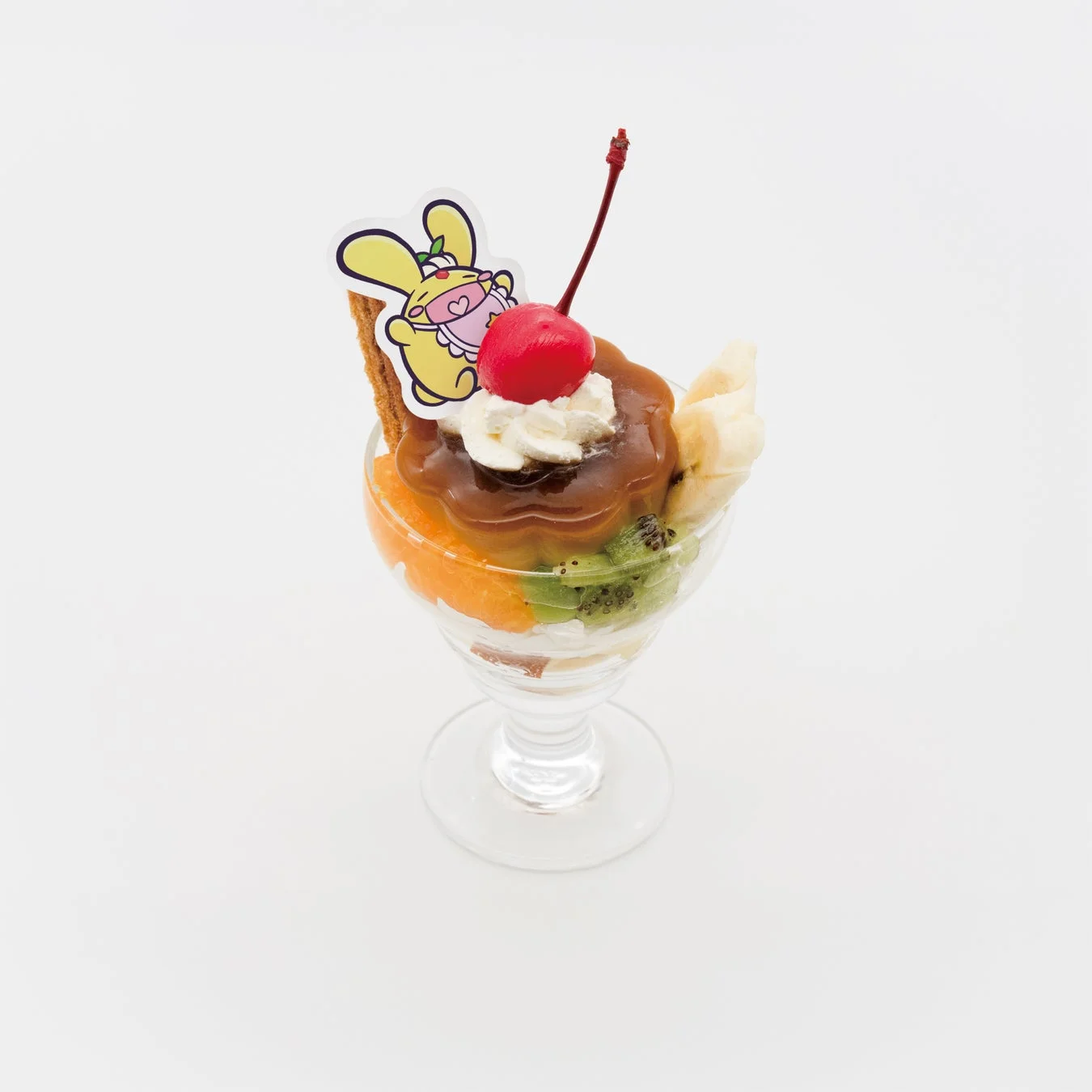 白い背景に、カラメルプリン、ホイップクリーム、様々なフルーツ（チェリー、バナナ、キウイ、オレンジ）が層になったパフェが写っています。かわいいウサギのキャラクターの飾りも添えられた、色鮮やかなデザートです。