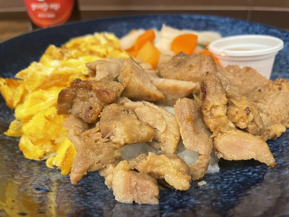 青い皿に盛り付けられた、ご飯の上に炒めた肉とスクランブルエッグがのった食欲をそそる一皿です。付け合わせには、人参や大根のようなピクルスと、小さなカップに入ったソースが添えられています。