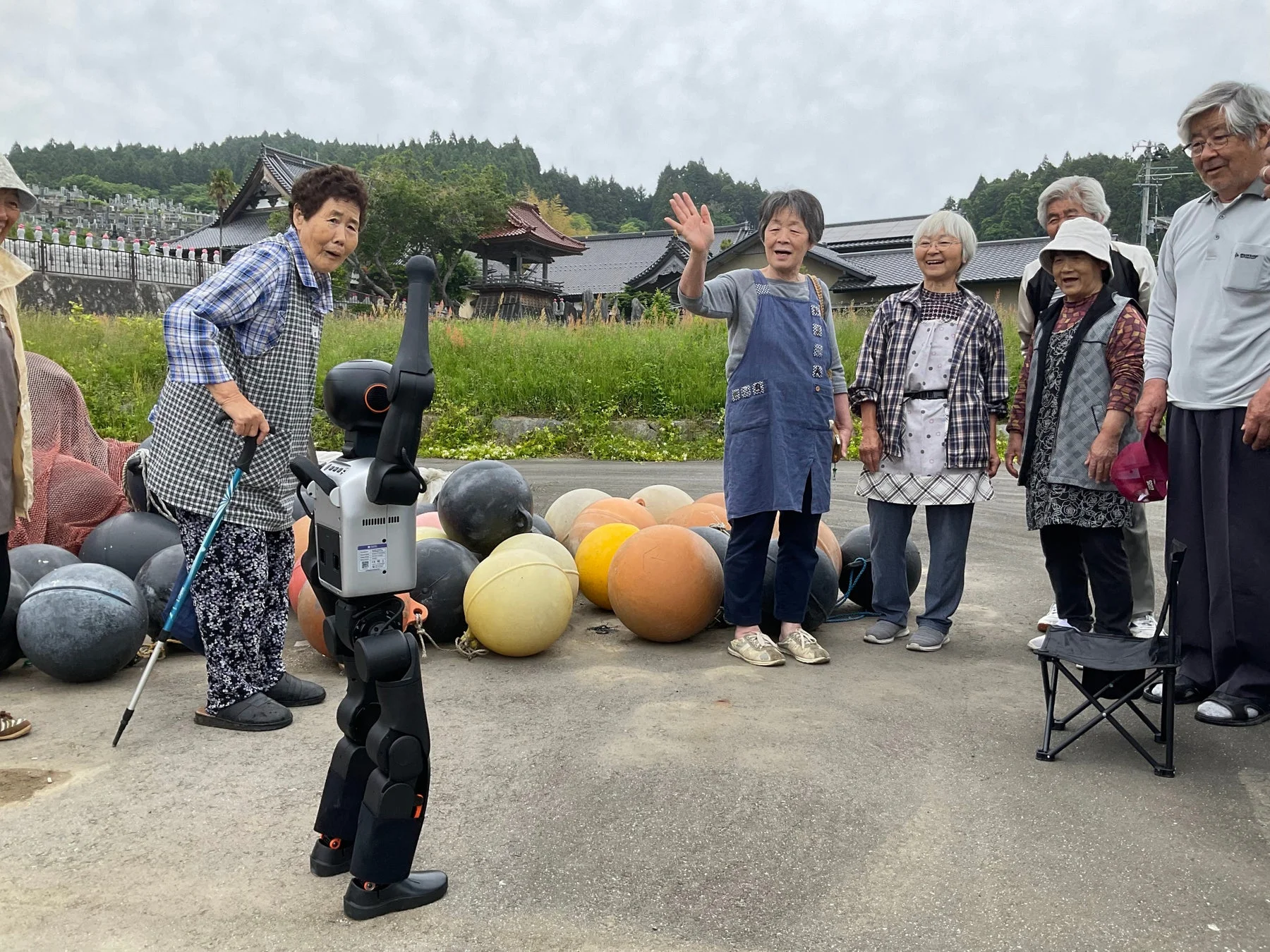 高齢者たちが屋外でロボットと交流している様子を捉えた写真です。背景には様々な色のブイが積まれ、遠くには日本の伝統的な建物や墓地が見えます。参加者たちは笑顔で、和やかな雰囲気が感じられます。