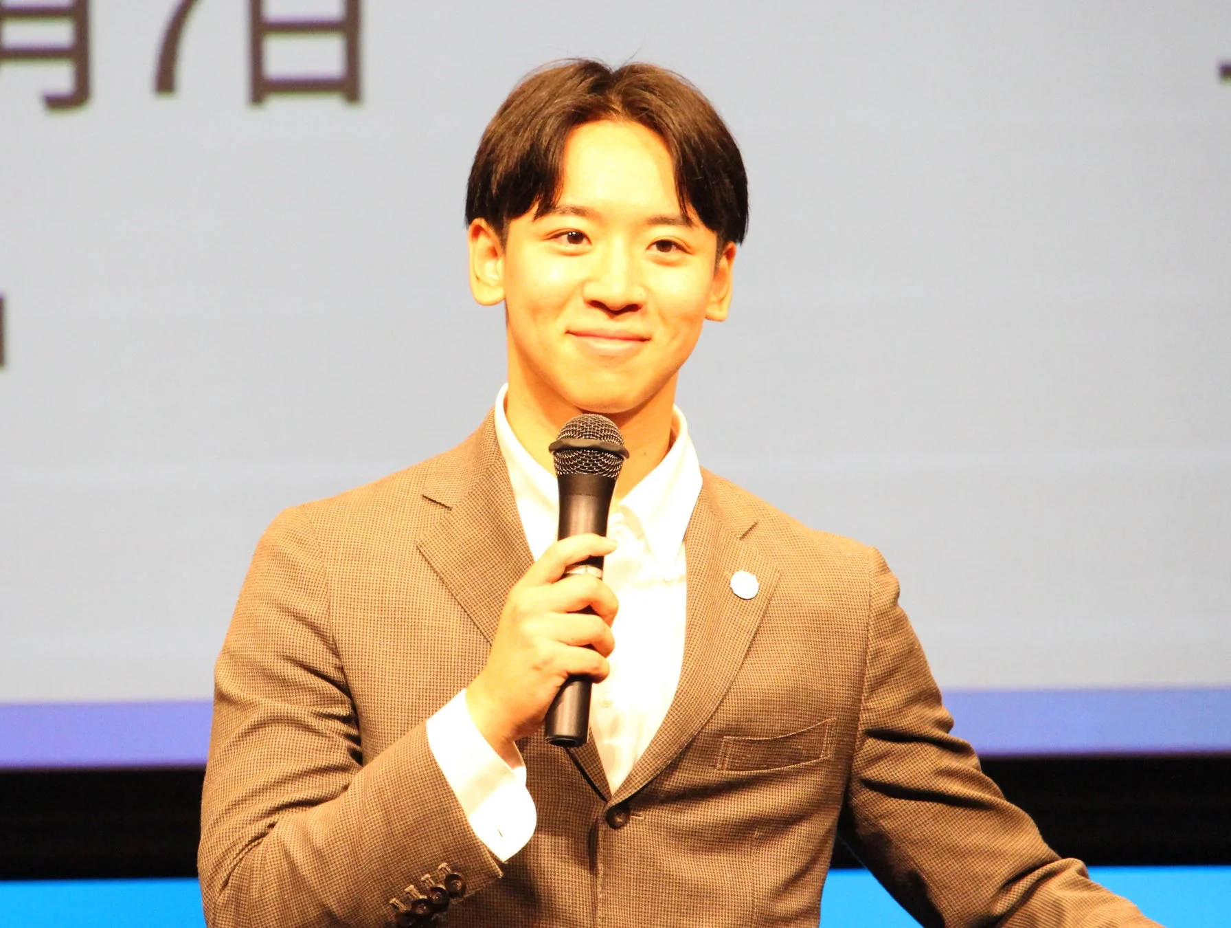 スーツを着た若い日本人男性がマイクを持って笑顔で話している様子。