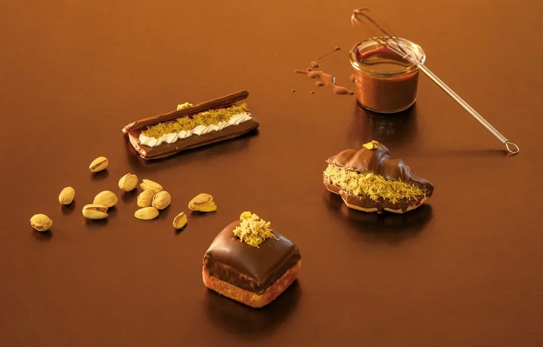 チョコレートでコーティングされた様々なデザートが、ピスタチオと共に茶色の背景に配置されています。チョコレートソースとホイッパーも添えられ、美味しそうなスイーツの魅力が伝わる一枚です。