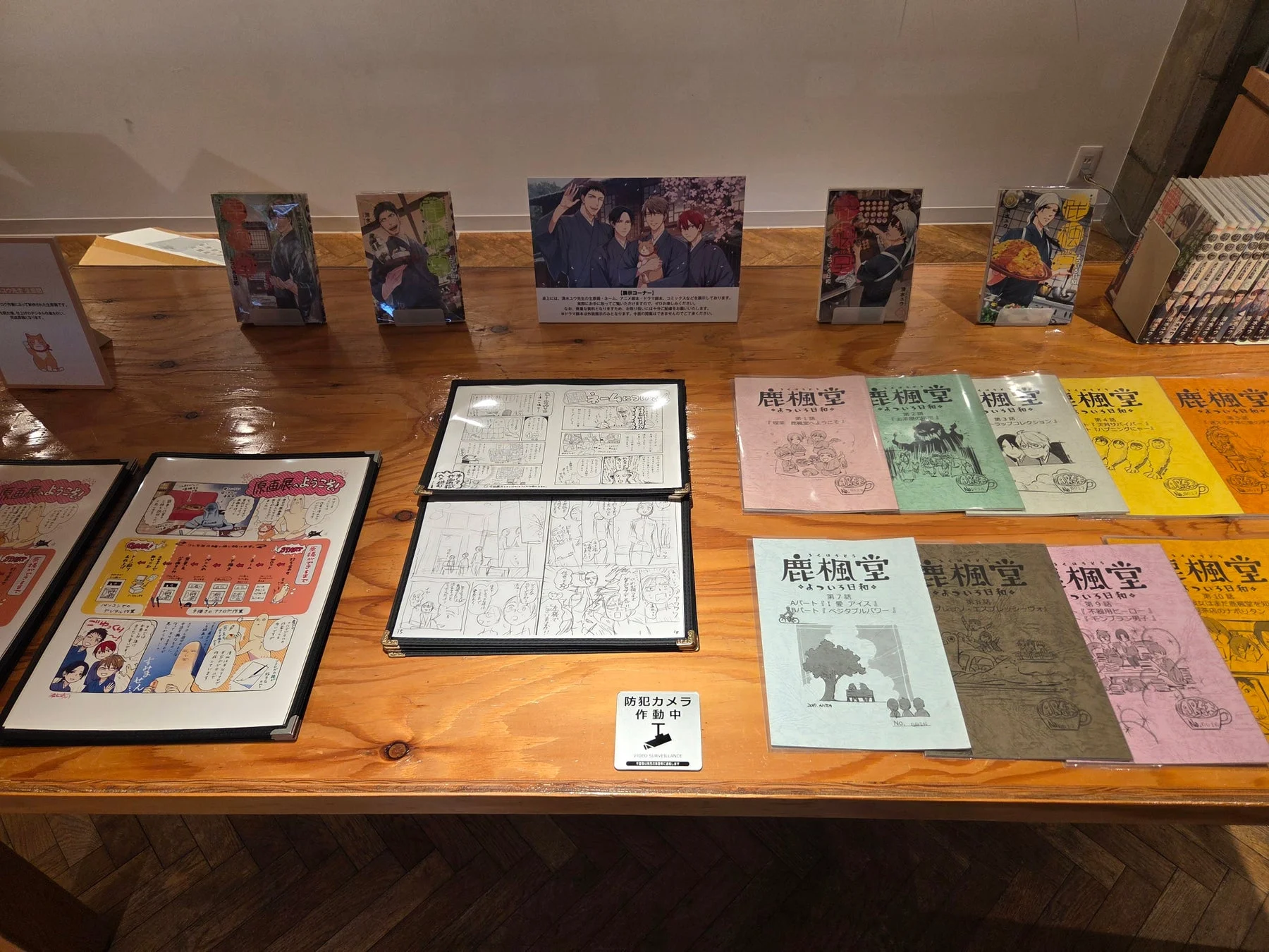 漫画「鹿楓堂よついろ日和」の展示会の様子を捉えた画像。清水ユウ先生によるアニメ関連グッズ、ドラマCD、コミックスなどが展示されており、複製原画やキャラクターのイラストも並んでいる。作品の世界観が伝わるイベント会場の風景。