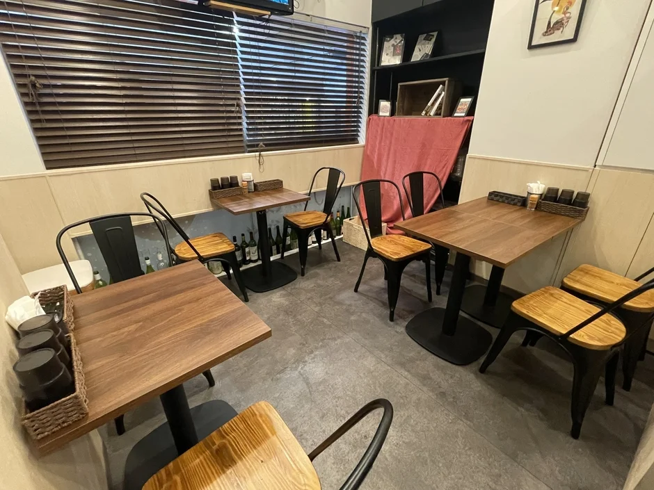 複数の木製テーブルと金属製の椅子が並べられた、カジュアルな雰囲気の飲食店内の様子です。窓にはブラインドがあり、壁際には棚が設置されています。