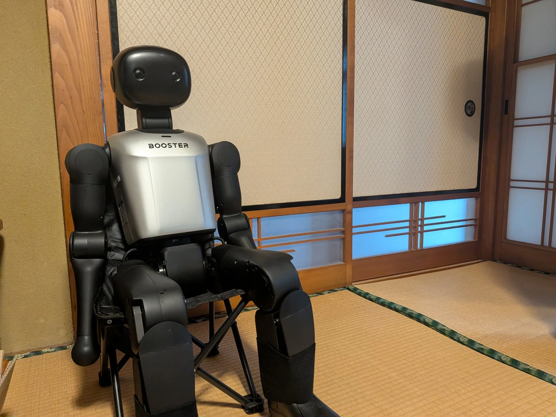 畳敷きの和室で、人型ロボットが小さな椅子に座っている様子です。背景には伝統的な模様の障子があり、ロボットの胸には「BOOSTER」の文字が見えます。