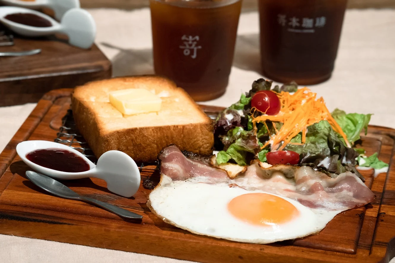 トーストにバター、目玉焼きとベーコン、サラダが盛り付けられた洋風の朝食プレート。奥にはアイスコーヒーが置かれ、ジャムも添えられています。