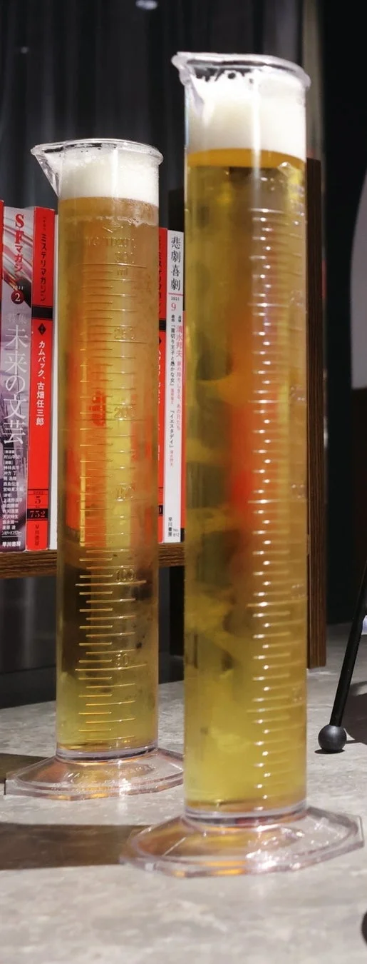 本棚を背景に、メスシリンダーのような容器に泡立つビールが2杯置かれている