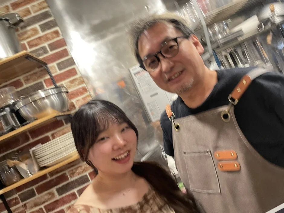 厨房のような場所で、エプロンを着用した男性と女性が笑顔でカメラに写っています