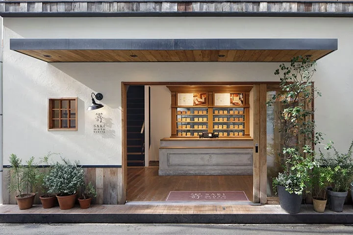 モダンなデザインのパン屋「嵜本 SAKI moto bakery」の店舗外観。木材と白壁が調和し、店頭には緑の鉢植えが並んでいます。店内には焼き菓子が並べられたショーケースが見え、温かい雰囲気が伝わります。