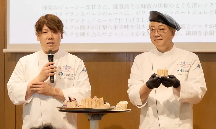 シェフコート姿の2人の男性パティシエがイベントでスイーツを紹介しています。左の男性がマイクで話し、右の男性がケーキを提示。テーブルには美しいデザートが並び、背景には洋梨や蜂蜜を使ったデザートの説明文が映し出されています。