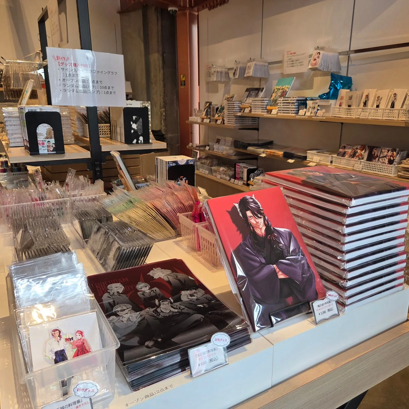 「魔王城の料理番」のグッズ販売コーナー。クリアファイルや書籍など様々な商品が整然と並べられ、購入制限や価格が提示されている。