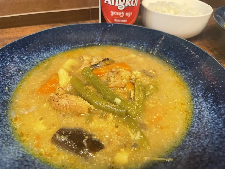 鶏肉、インゲン、人参、ナスなどが入った黄色いスープまたはカレーが青い器に盛られています。奥には白いご飯と「Angkol」と書かれた赤いラベルのボトルが見える、アジアンテイストの食事風景です。