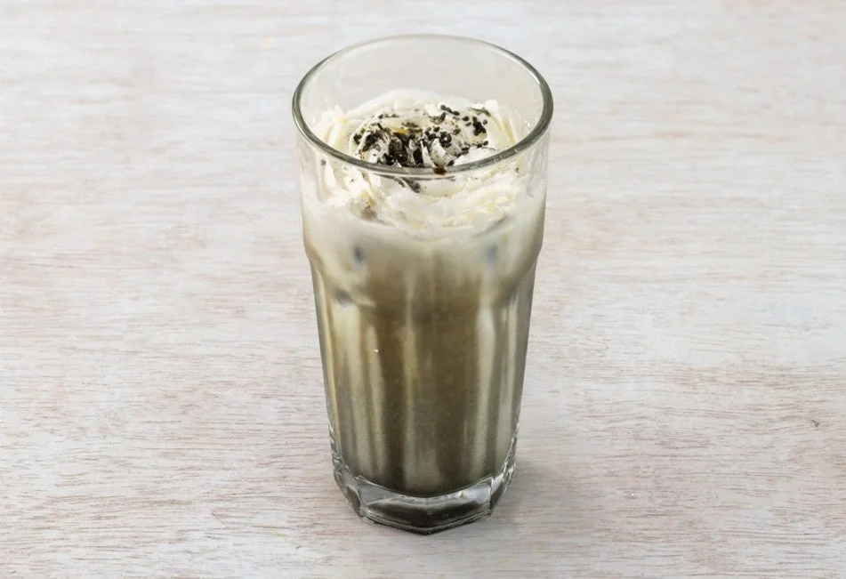 ホイップクリームと黒い粒がトッピングされた、背の高いグラスに入った冷たい飲み物の写真。淡い木目のテーブルに置かれています。