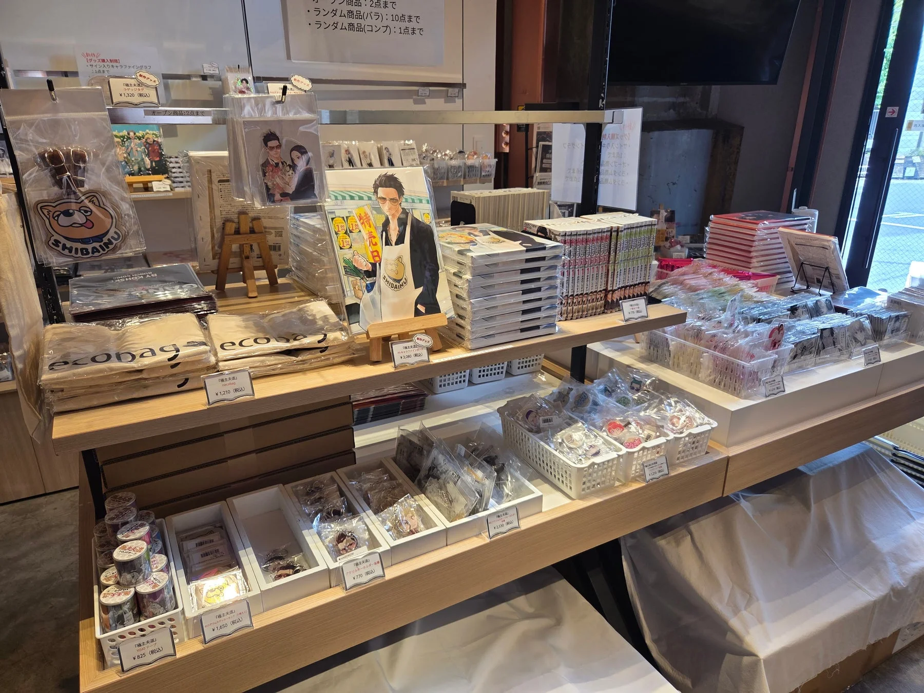 人気漫画「極主夫道」の公式グッズ販売店の様子です。エコバッグ、キーホルダー、コミックス、アクリルスタンドなど、様々な商品が木製棚に陳列され、価格や購入制限が示されています。
