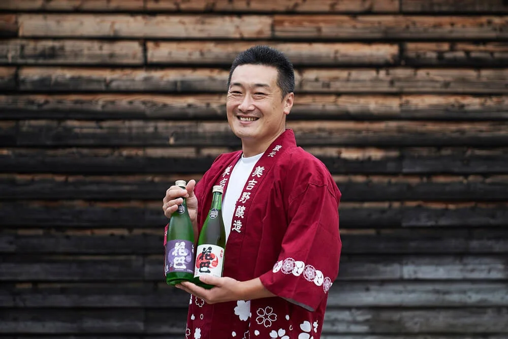 赤い法被を着た笑顔の男性が美吉野醸造の日本酒「花巴」の瓶を手に持つ