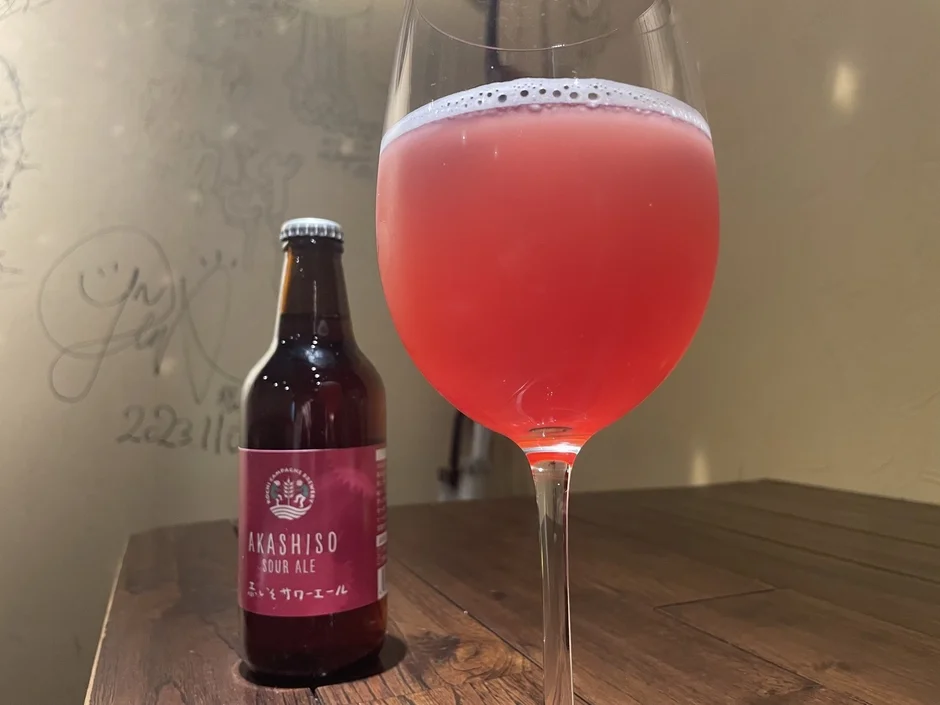 木製のテーブルの上に、泡の立った赤紫色の飲み物が入ったグラスと「AKASHISO SOUR ALE」と書かれた瓶が置かれています。背景には手書きの文字が見える壁があります。