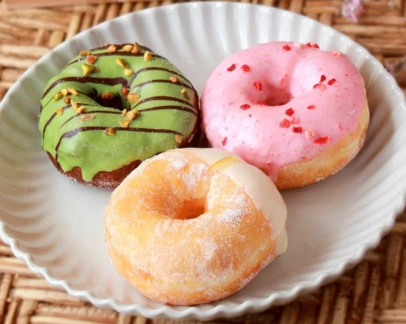 白い皿に3つの異なるフレーバーのドーナツが盛り付けられています。緑色の抹茶風味、ピンク色のストロベリー風味、そしてシュガーパウダーのかかったプレーンなドーナツがあり、美味しそうなデザートの光景です。
