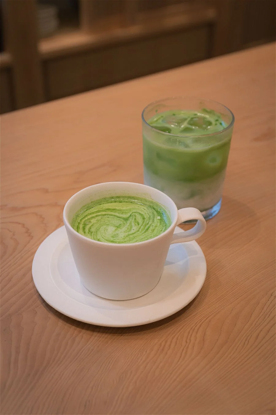 木製のテーブルに置かれた温かい抹茶ラテとアイス抹茶ラテ。鮮やかな緑色のドリンクが心地よい休憩時間を提供している様子です。