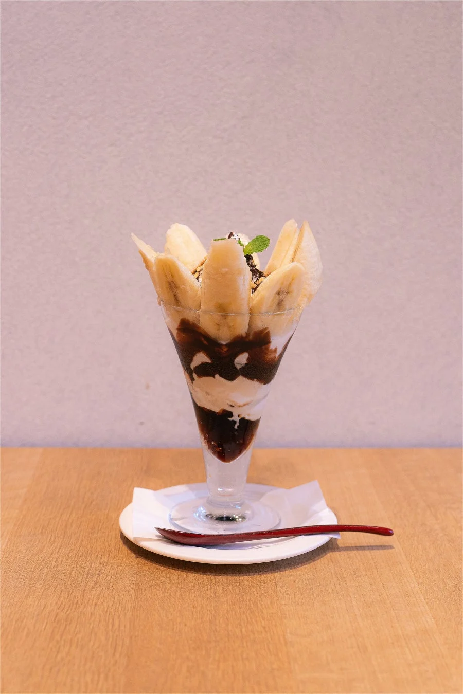 スライスされたバナナがグラスの周りに飾られ、チョコレートソースとホイップクリームが層になったバナナパフェが木製のテーブルに置かれています。ミントの葉とナッツがトッピングされており、白い皿と赤いスプーンが添えられています。