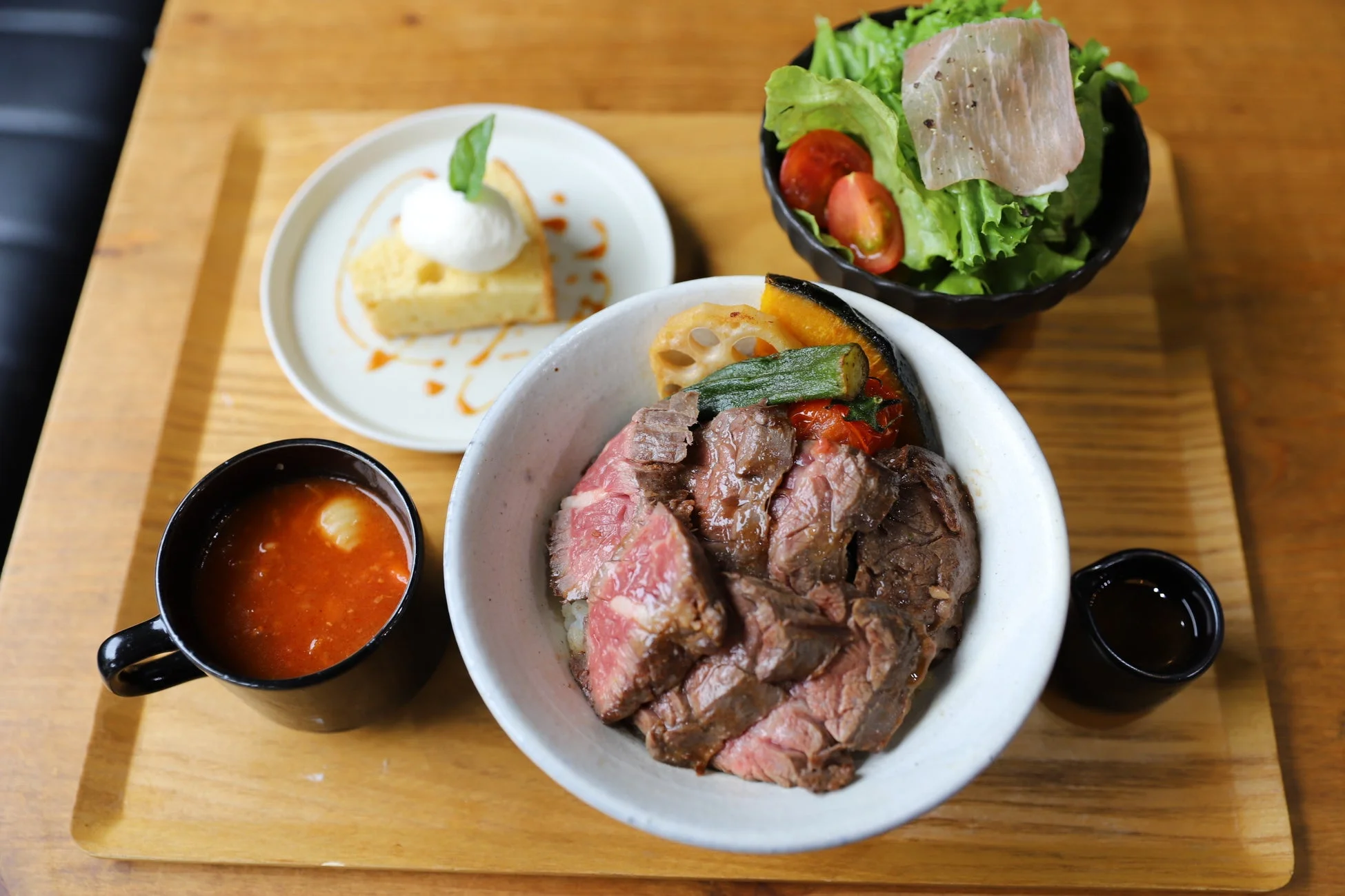木製トレーに盛り付けられたランチセット。メインはローストビーフ丼で、彩り豊かな焼き野菜が添えられています。サイドには生ハムサラダ、トマトスープ、そして食後のデザートとしてケーキも含まれており、充実した食事が楽しめます。