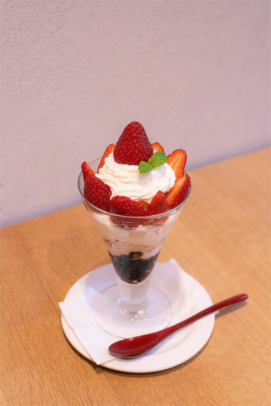 新鮮ないちごがたっぷり飾られた、白い生クリームとミントの葉が美しいパフェです。透明なグラスに入っており、層になった具材が見えます。木製のテーブルに置かれた白い皿の上には赤いスプーンが添えられています。