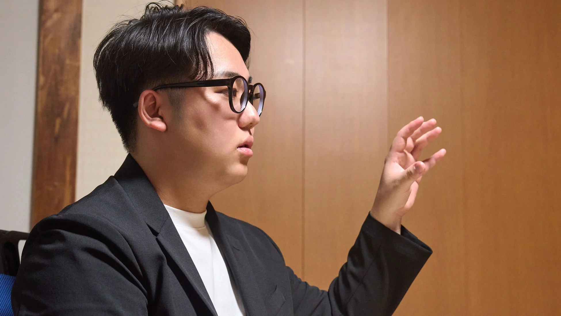 眼鏡をかけた若い男性が黒いジャケットと白いシャツを着用し、右手を上げて何かを説明しているような様子