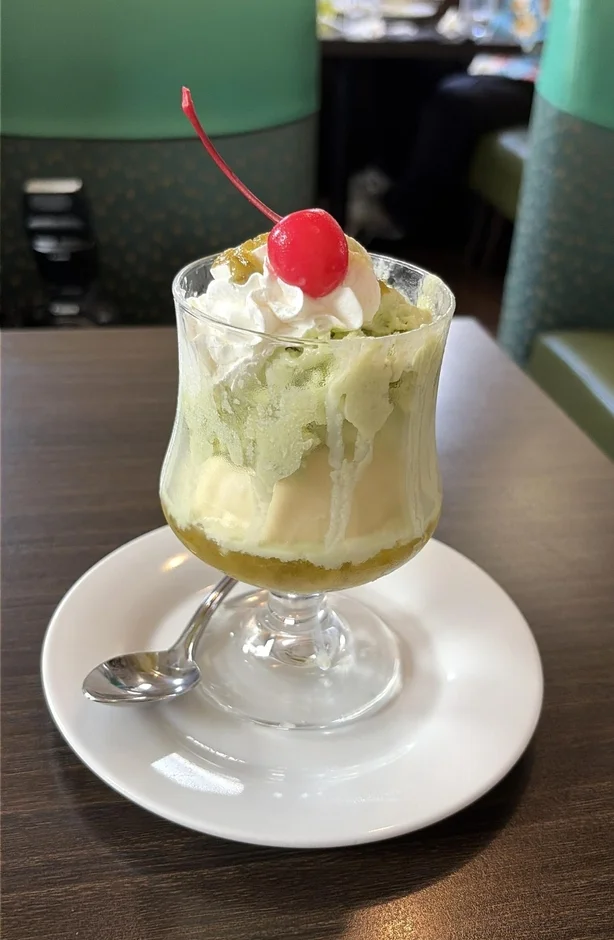 グラスに入った抹茶色のパフェで、上にはホイップクリームと赤いサクランボが飾られています。カフェのような場所で提供されている美味しそうなデザートです。