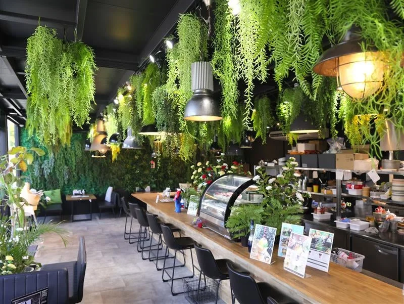 緑豊かな植物が天井や壁を飾り、自然を感じさせるモダンなカフェの店内風景。長い木製カウンターにはバーチェアが並び、ショーケースやメニューが見える、落ち着いた雰囲気の空間。