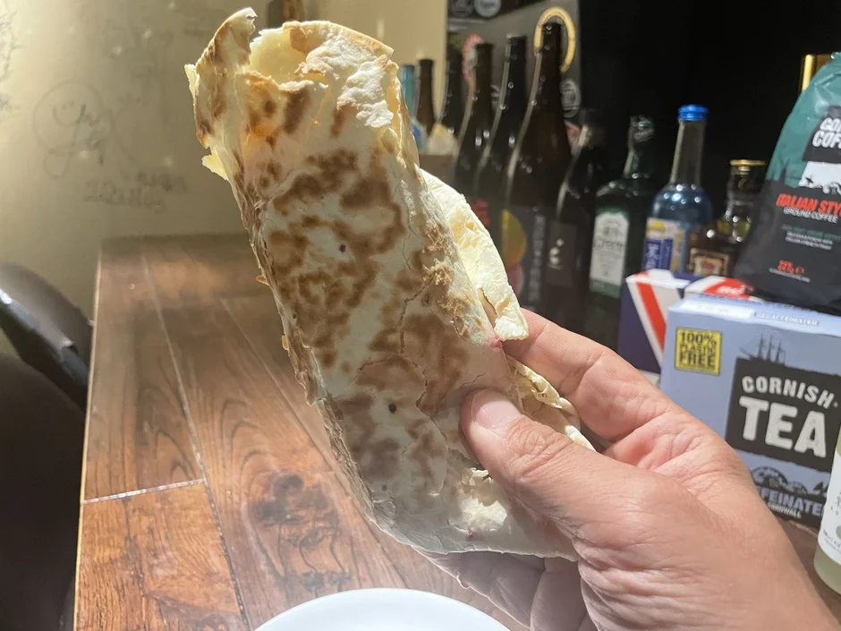手がラップサンドのような食べ物を持っており、背景にはコーヒー豆の袋や紅茶の箱、様々な瓶が置かれた木製のテーブルが写っています。