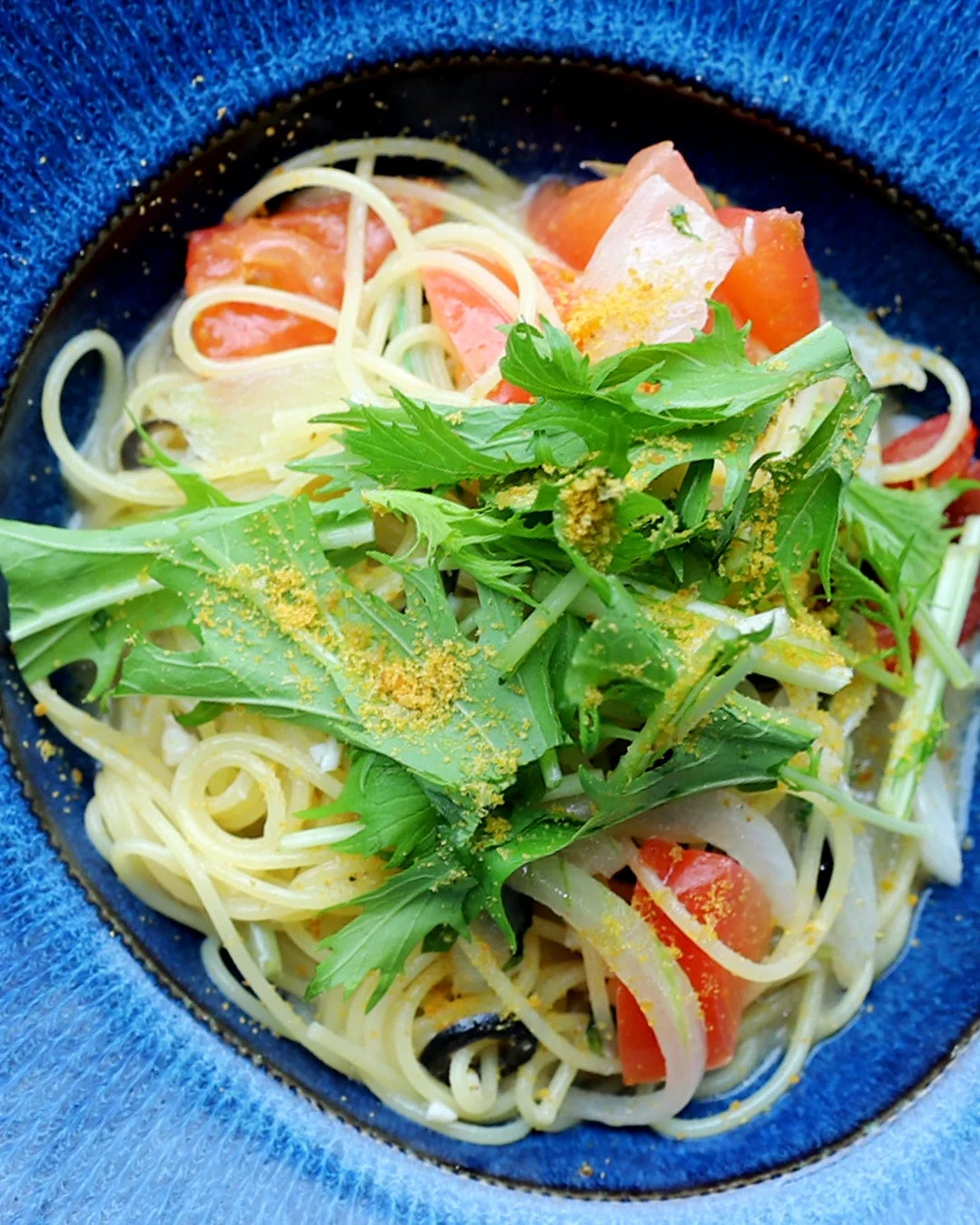 青い器に盛り付けられた、トマト、玉ねぎ、水菜などの野菜がたっぷり入ったスパゲッティ。黄色い粉末が振りかけられており、彩り豊かでヘルシーな印象の料理です。
