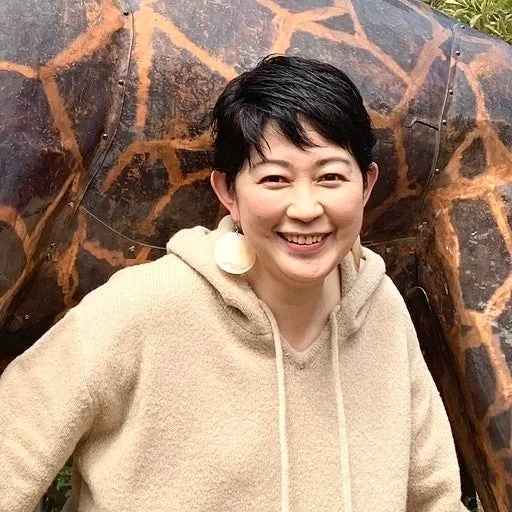 ショートヘアのアジア人女性が、ベージュのパーカーを着て、大きな丸いイヤリングをつけ、明るい笑顔でカメラを見ている。背景には、茶色と黒の模様があるオブジェが写っている。