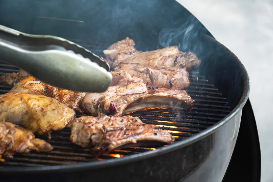 炭火のグリルで骨付き肉や鶏肉が焼かれている様子を捉えた画像です。煙が立ち上り、トングで肉をひっくり返している最中のようで、活気あるバーベキューシーンが伝わってきます。