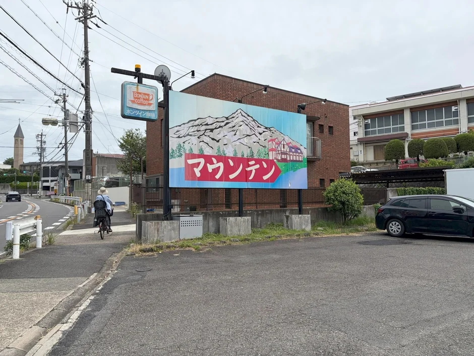 「マウンテン」と書かれた大きな看板が目立つ、日本の街角の風景。隣には「BONTAIN coffee」の小さな看板があり、喫茶店の存在を示唆している。道路を自転車で走る人が見え、遠くには教会の尖塔も確認できる。全体的に曇り空で、日常的な情景が広がっている。
